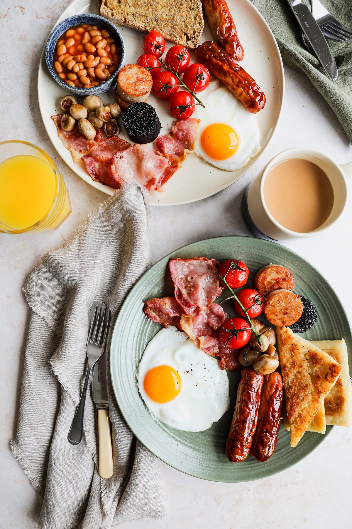 Two plates of Irish breakfast with sausages, rashers, eggs, puddings and tomatoes, with tea and orange juice